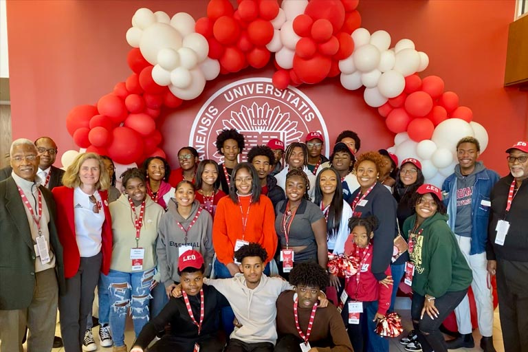 A group of individuals in various attire, including formal and casual wear, as well as university merchandise, stand in front of a red wall. The scene is festively decorated with red and white balloons, and some attendees wear lanyards, from a university event.